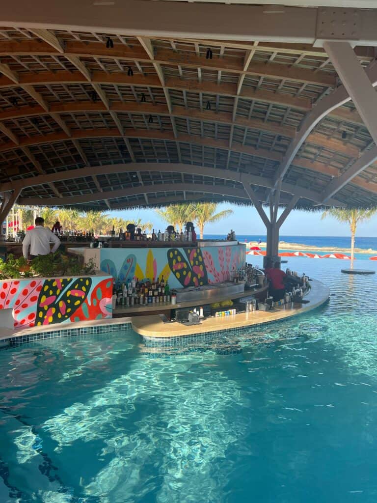 A wide-angle view of a vibrant, curved swim-up pool bar sheltered under a massive wooden, grid-pattern pavilion roof with a thatched exterior. The front of the bar features colorful, tropical pop-art murals with bright floral and leaf designs. Patrons stand or sit along the counter, while a bartender in a red shirt works behind it. The sparkling blue pool water fills the foreground, leading out toward an open view of the ocean and distant palm trees under a clear sky, highlighting a relaxing resort amenity booked through straight away travel.