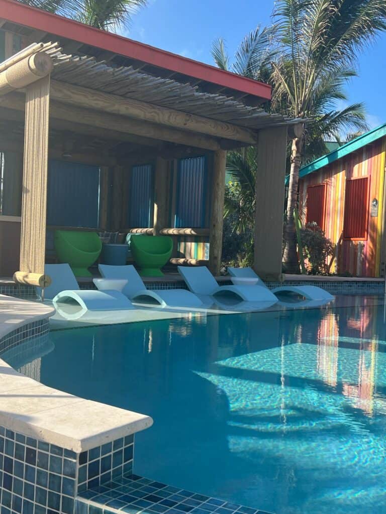 A tropical resort swimming pool with light blue in-water lounge chairs lined up on a shallow ledge. In the background, a wooden cabana with a thatched trim roof features bright green accent chairs and blue privacy curtains. To the right, a colorful, striped building and tall palm trees stand under a clear blue sky, showcasing a premium relaxation spot arranged by straight away travel.