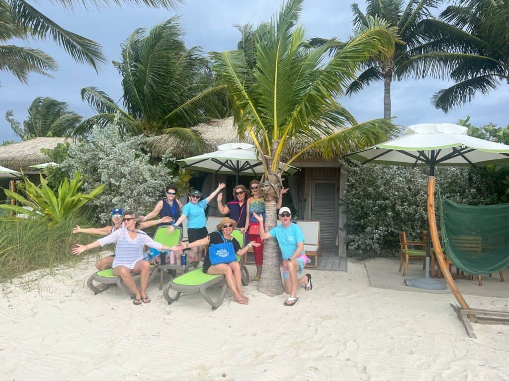 A group of travelers on a Straight Away Travel trip posing playfully on a sandy beach. The group is gathered around a vibrant palm tree in front of a tropical resort hut with lounge chairs and beach umbrellas. They are smiling and waving at the camera, capturing a fun vacation moment under a slightly overcast sky.