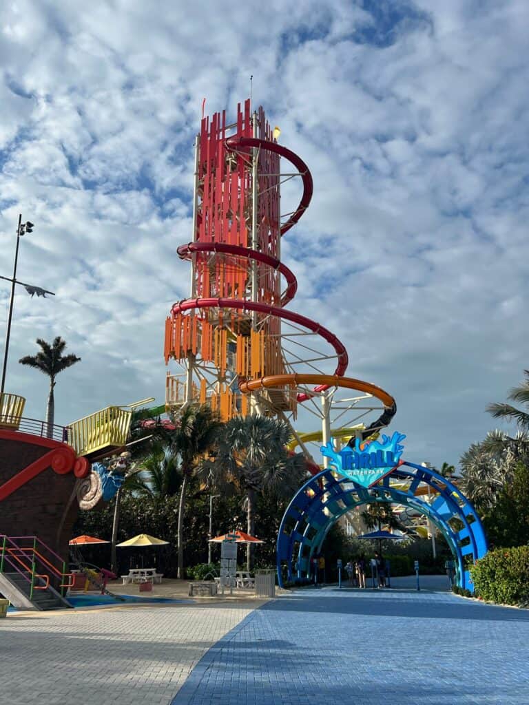 A Straight Away Travel vacation destination featuring Daredevil's Tower at Perfect Day at CocoCay. The massive, vibrant orange and red water slide tower dominates the landscape under a partly cloudy sky. In the foreground, a blue brick pathway leads toward a bright blue arched entrance labeled "Thrill Waterpark," with a pirate ship-themed play area visible on the left.