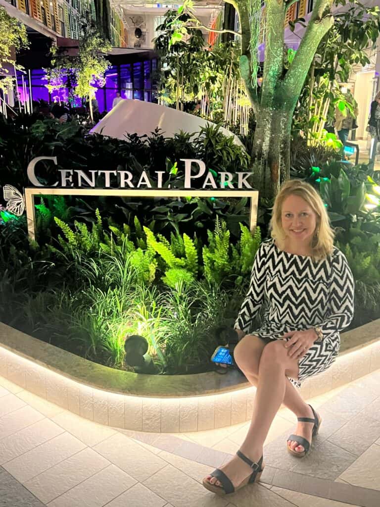 A smiling blonde woman sits on the edge of a lighted planter box in the Central Park neighborhood of a Royal Caribbean cruise ship. She is wearing a black-and-white chevron pattern dress and dark wedge sandals while holding a phone. Behind her, a sign reads "Central Park" amidst lush green ferns, tropical plants, and a large tree lit with green ambient lighting, capturing an evening cruise experience booked through straight away travel.