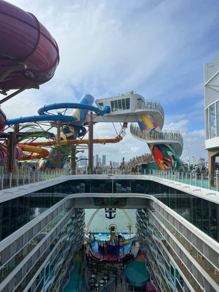 A high-angle view looking down into the open-air central boardwalk of a large cruise ship, framed by multi-story balcony cabins on both sides. Towering above the upper deck are massive, colorful water slides, including a prominent winding blue and yellow slide structure and a large purple tube slide on the left. In the lower center, a children's splash zone and seating area are visible, leading toward an open view of the ocean and a distant city skyline under a partly cloudy sky, showcasing an action-packed family vacation experience booked through straight away travel.