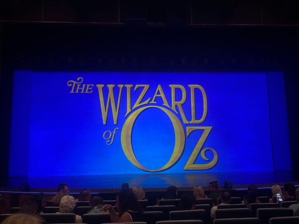 A Straight Away Travel theater excursion showcasing a stage production of The Wizard of Oz. The bright blue stage curtain features the iconic "The Wizard of Oz" title in large, gold, stylized lettering. In the foreground, the dark silhouettes of the audience members can be seen seated in theater rows, waiting for the performance to begin.