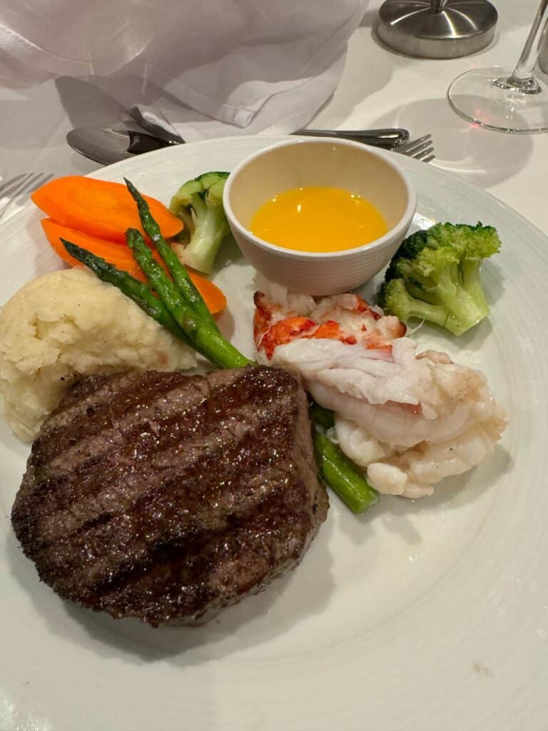 Alt Text: A gourmet surf and turf dinner enjoyed on a Straight Away Travel vacation. A white plate features a perfectly grilled steak with visible sear marks, a tender lobster tail, and a scoop of mashed potatoes. The meal is accompanied by steamed vegetables, including bright orange carrots, green broccoli florets, and asparagus stalks, along with a small ceramic bowl of drawn melted butter.