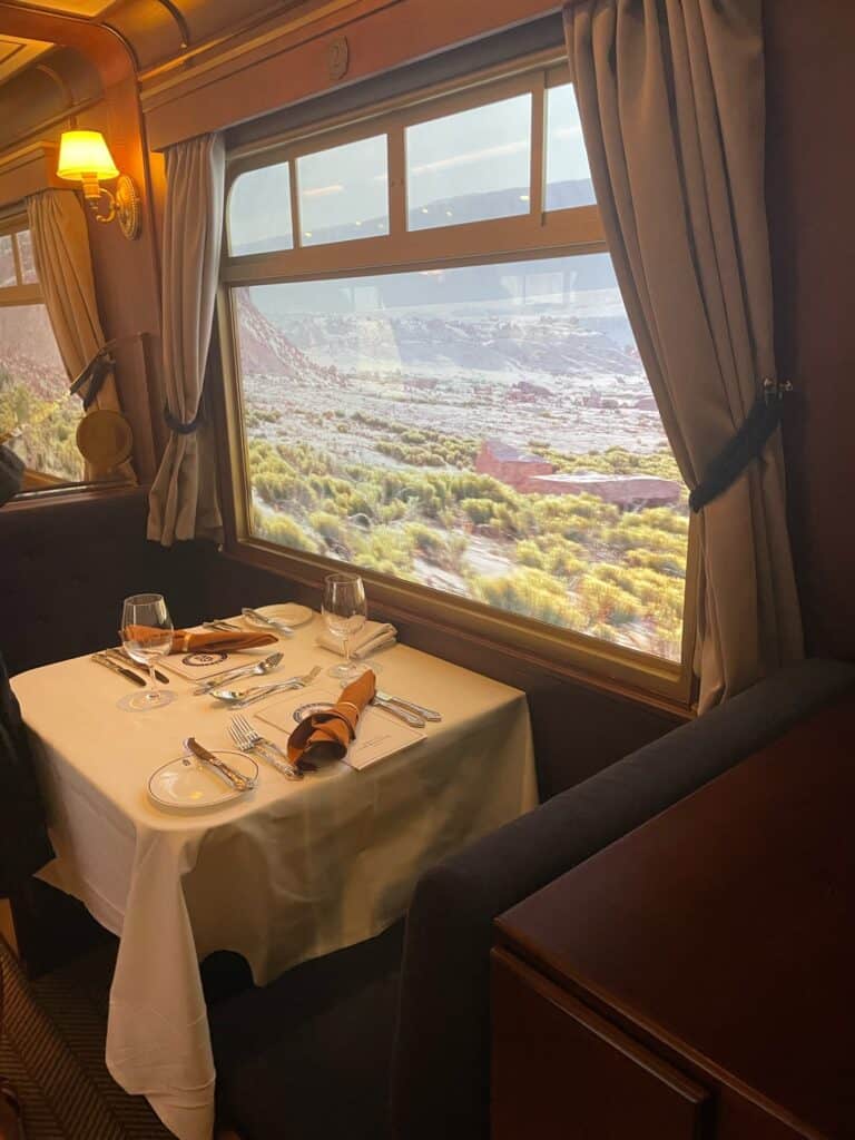 A luxury rail dining experience booked through Straight Away Travel. The interior of an elegant train dining car features a beautifully set table with a crisp white tablecloth, fine silverware, folded orange napkins, and wine glasses next to a plush booth seat. A large window draped with neutral curtains offers a scenic view of a passing desert landscape with low shrubs and distant rocky formations under a warm, ambient light.