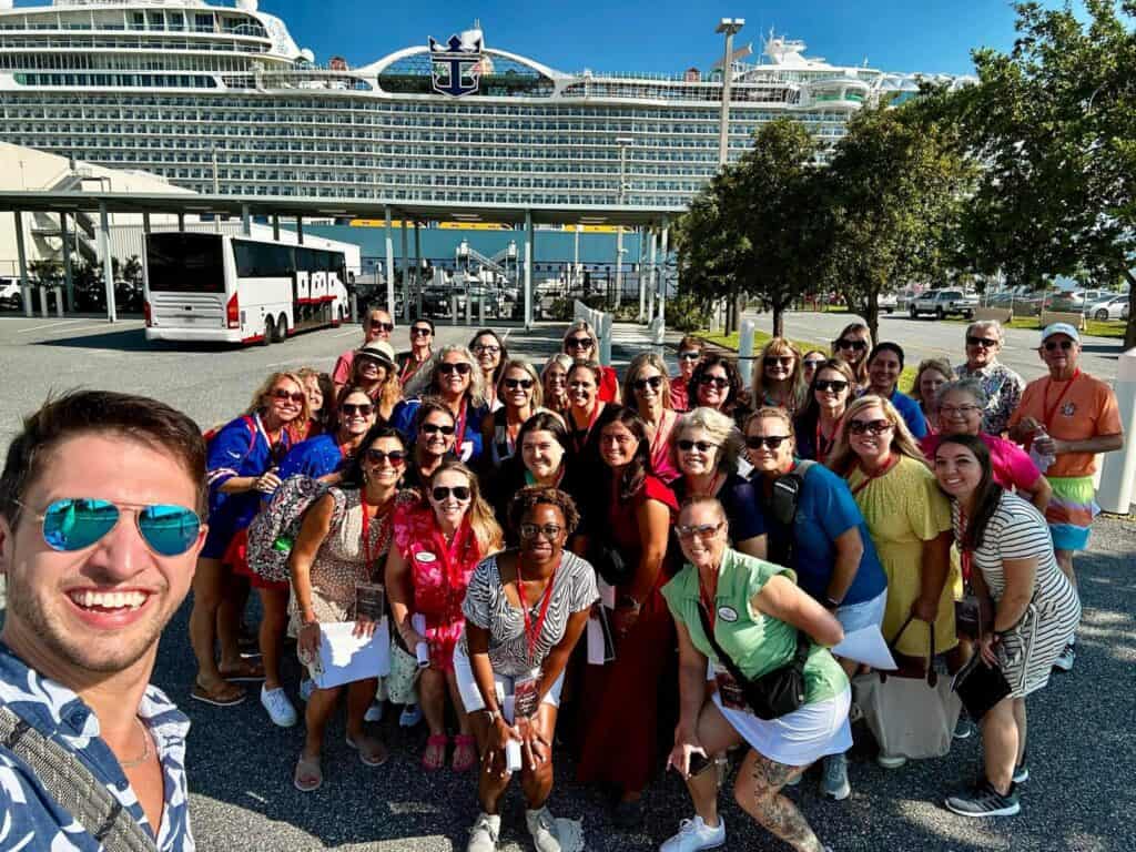 A large group of Straight Away Travel clients and agents smiling for a group selfie at a cruise port terminal. In the foreground, a man in sunglasses smiles widely as he takes the photo, capturing a large, diverse group behind him on the asphalt. In the background, a massive Royal Caribbean cruise ship looms large under a bright, sunny sky, alongside a white tour bus.