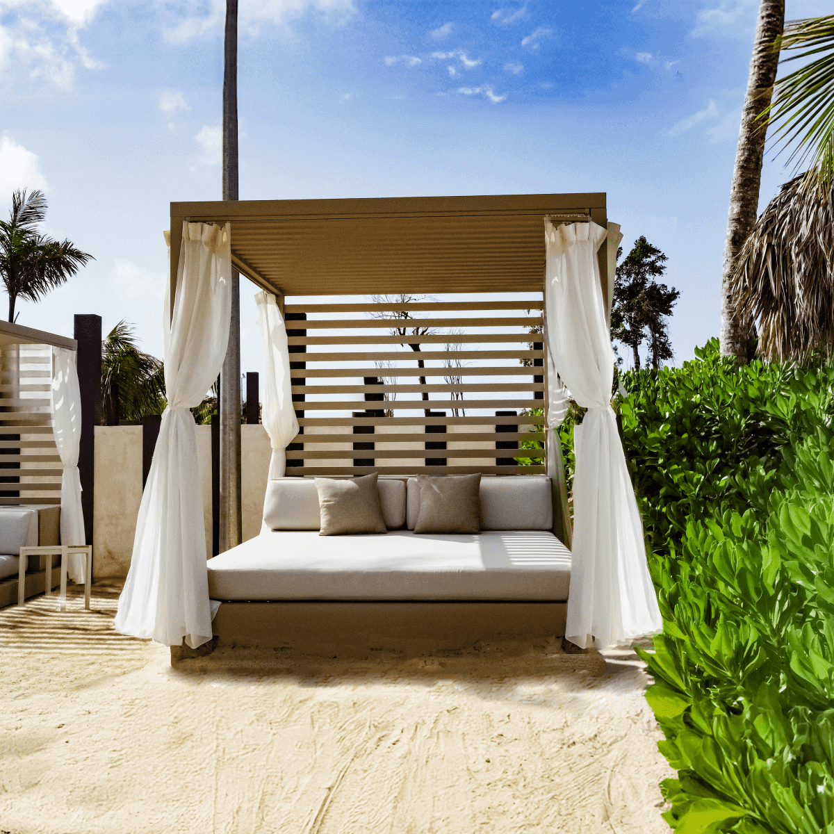 A modern, square luxury beach cabana set on fine sand, featuring a beige frame with a slatted back wall for partial shade. The cabana is outfitted with a plush white mattress bed, matching back cushions, two tan throw pillows, and flowing white privacy curtains tied to the corner posts. Lush green bushes flank the right side, while palm trees and a bright blue sky with light clouds appear in the background, showcasing a premium relaxation spot for a straight away travel itinerary.