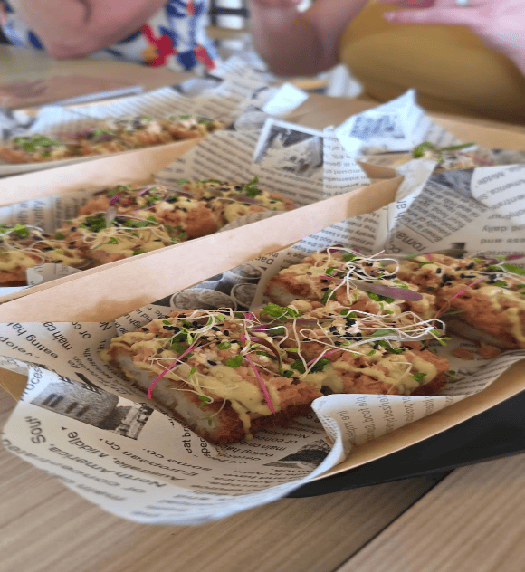 A close-up, shallow-depth-of-field shot of several portions of a gourmet appetizer served on a table. The dish features a crispy, fried base topped with a savory spread, drizzled with a light green sauce, and garnished with black sesame seeds, fresh microgreens, and delicate sprouts. The food is neatly presented on black trays lined with decorative newspaper-print wax paper. In the blurred background, people are seated at the table enjoying the dining experience, showcasing a unique culinary highlight from a straight away travel excursion.