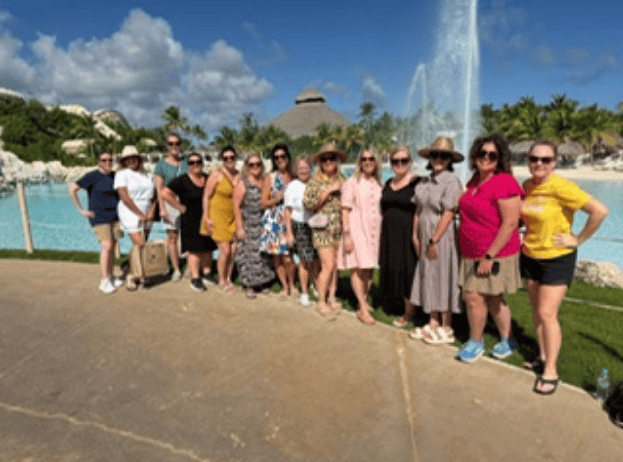 A group of fourteen women posing together in a line on a paved pathway at a tropical resort or water park. They are dressed in casual summer attire, dresses, and sun hats, with many wearing sunglasses. Behind them is a large, light-blue pool or lagoon with a prominent water fountain spraying high into the air, palm trees, and a large thatch-roofed palapa building under a blue sky with fluffy white clouds, capturing a group getaway organized by straight away travel.