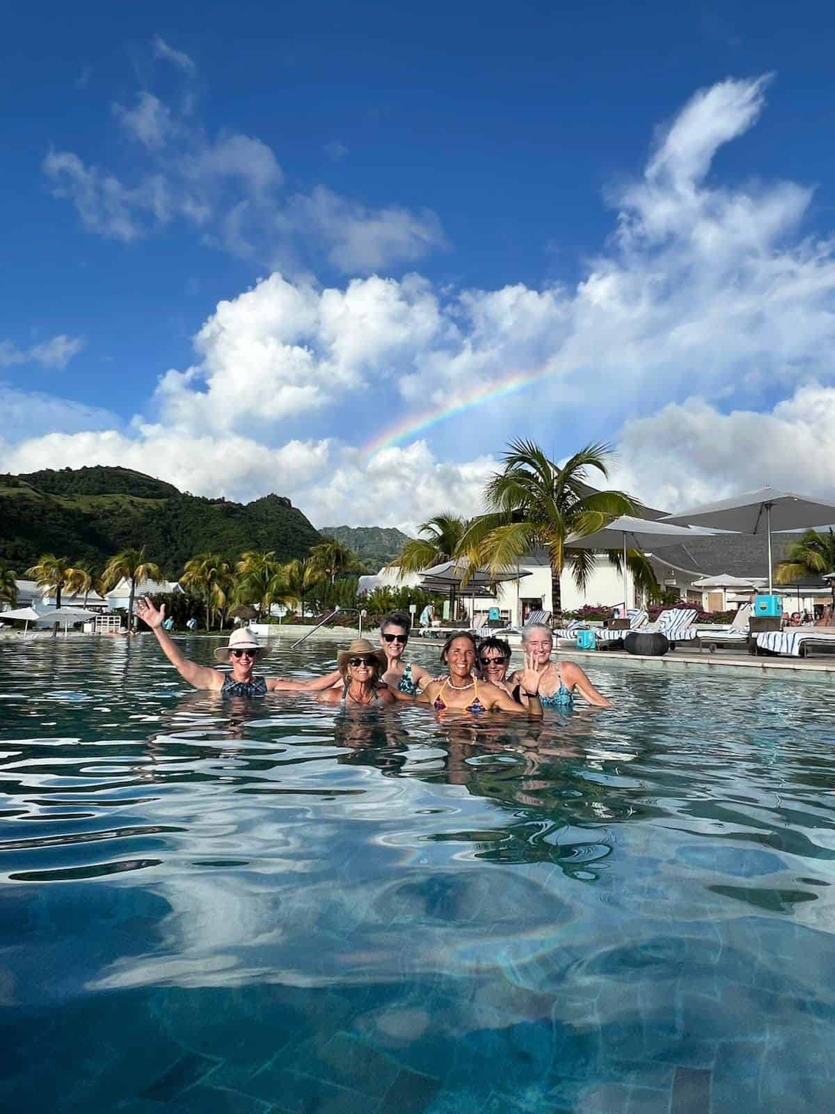 A tropical vacation experience curated by Straight Away Travel shows a group of six women smiling and wading in a large, clear resort swimming pool. They are gathered close together in the water, with some waving toward the camera. The luxurious backdrop features palm trees, white lounge chairs with umbrellas, and a lush green mountain range under a blue sky filled with dramatic white clouds and a vibrant rainbow cutting through the center.