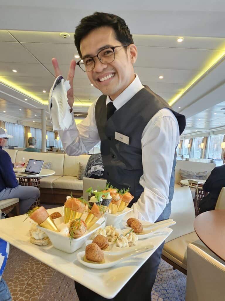 A smiling male server wearing glasses, a white dress shirt, a black tie, and a dark grey vest holds a large platter of gourmet appetizers. He flashes a peace sign with one hand while holding a white napkin. The platter features small savory cones filled with salmon and dill, alongside single-bite items served on white ceramic spoons. The bright, upscale background shows a cruise ship lounge with cream-colored booths and tables, highlighting the premium hospitality and dining experiences offered by straight away travel.