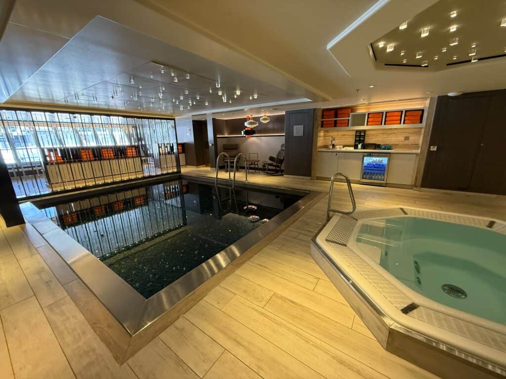 An interior view of a tranquil luxury spa and thermal suite area. The space features a small, dark-tiled plunge pool with a metallic rim on the left and a light blue octagonal hot tub or jacuzzi in the foreground on the right. The floor is made of light wood-style plank tiles. In the background, a refreshment station is stocked with towels and a small beverage cooler under soft, recessed lighting, showcasing a premium relaxation amenity on a cruise or resort vacation managed by straight away travel.