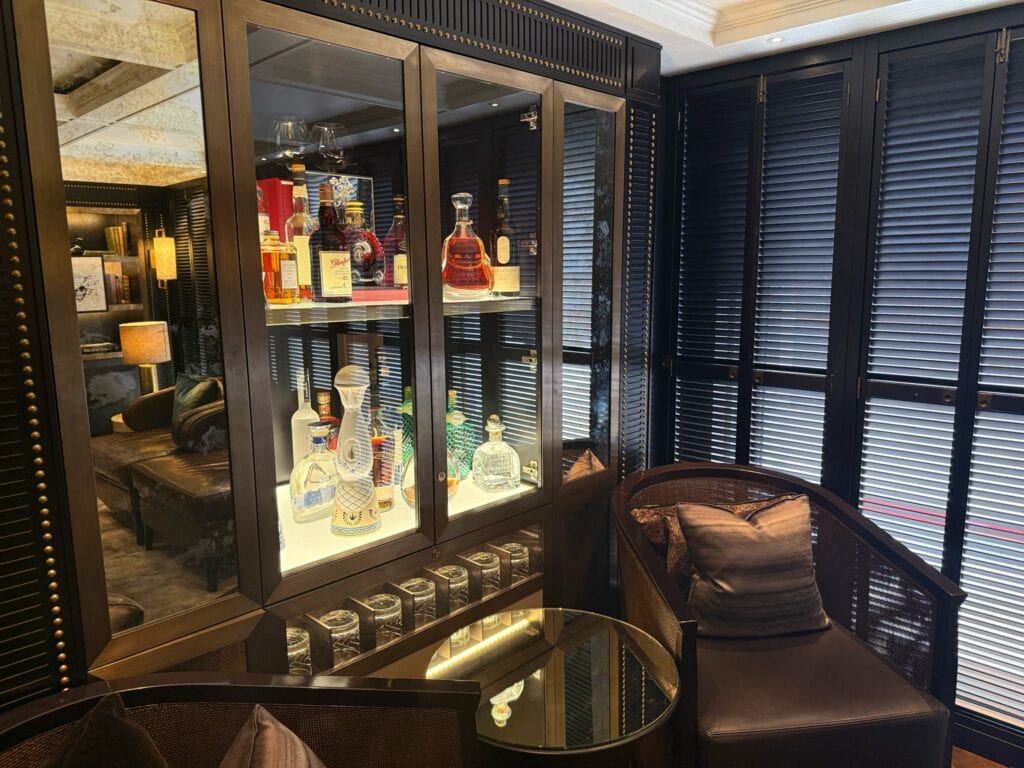 An interior view of a sophisticated, dark-paneled lounge or private bar featuring a lit glass display cabinet filled with premium liquor bottles, decanters, and glassware. To the right of the cabinet, a comfortable dark leather armchair with a decorative metallic-sheen pillow sits beside a small, round glass-top side table. The right wall is covered in elegant black louvered shutters, while a mirror on the left reflects an adjacent cozy seating area with a sofa and lamp, illustrating an exclusive onboard space perfect for a luxury itinerary by straight away travel.