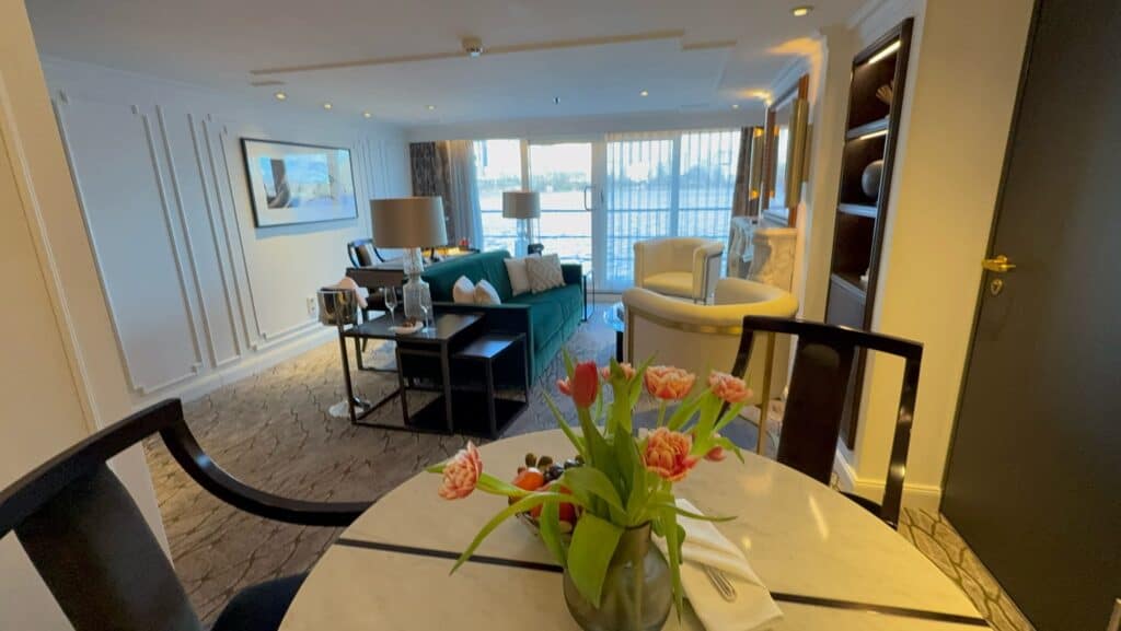 An interior view of a spacious luxury cruise ship suite or hotel room. In the foreground, a round dining table features a vase of pink and orange tulips. The background opens into a stylish living area furnished with a deep green sofa, two white club armchairs, and a small dining set next to wall molding details. Large floor-to-ceiling glass doors lead out to a private balcony overlooking the water, capturing a high-end accommodation option booked through straight away travel.