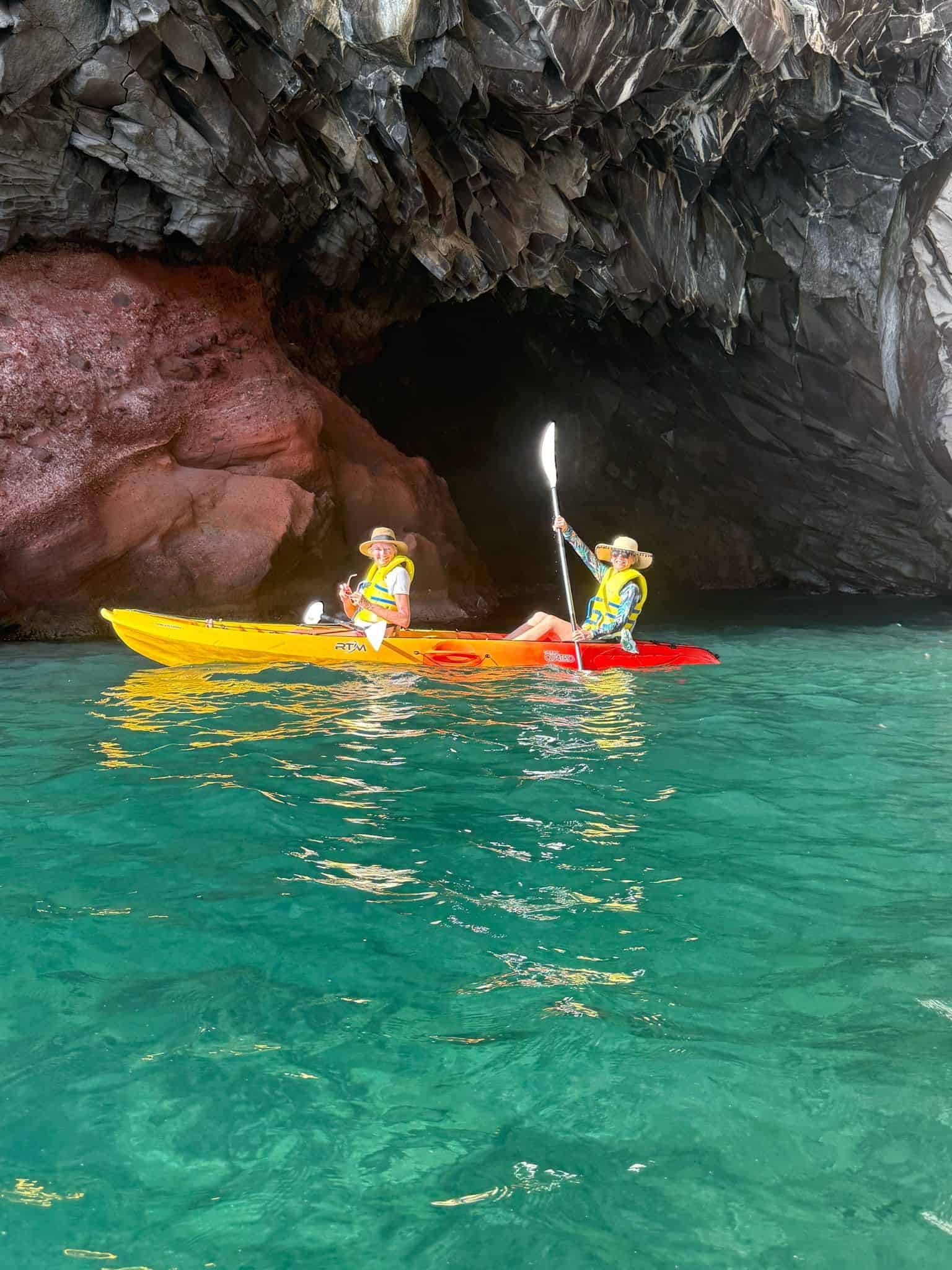 An outdoor adventure booked through Straight Away Travel features two people navigating a vibrant yellow and red tandem kayak on clear, turquoise water. Both kayakers are wearing yellow life vests and sun hats, with the person in the back holding a paddle aloft. They are positioned near the entrance of a dramatic sea cave, showcasing rugged, dark rock formations overhead and a smooth, reddish-brown rock face to the left.