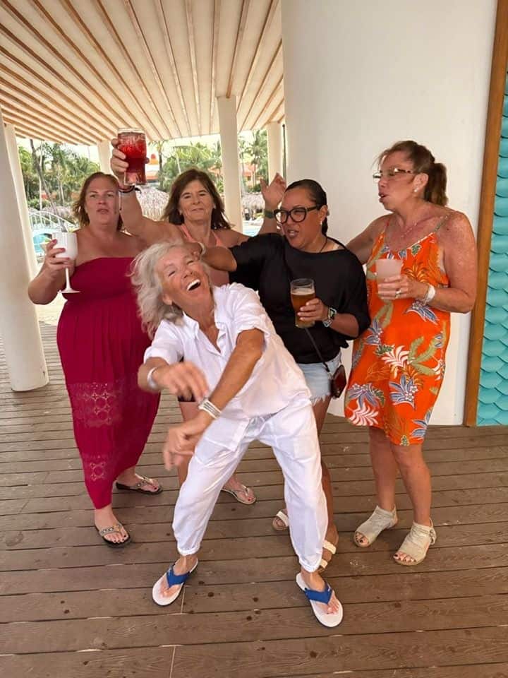 A lively vacation moment captured by Straight Away Travel shows a group of five women celebrating and dancing together under a shaded outdoor pavilion with a wooden deck. In the center, a woman with white hair dressed entirely in white laughs and dances energetically. Around her, the other women join in the fun, smiling, dancing, and holding up drinks, including cocktails and beers, in a festive resort atmosphere.