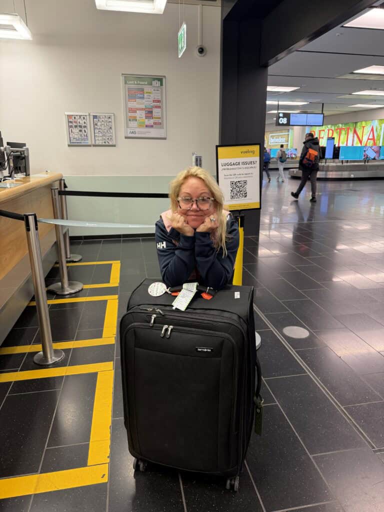 A blonde woman wearing glasses and a navy Helly Hansen jacket rests her chin in her hands, leaning on a large black Samsonite suitcase in an airport baggage claim area. She stands near a service counter with yellow floor markings and a yellow sign with a QR code titled "Luggage Issues?". In the background, a baggage carousel labeled with the number 08 is visible alongside other travelers, highlighting a common scenario managed smoothly by straight away travel.