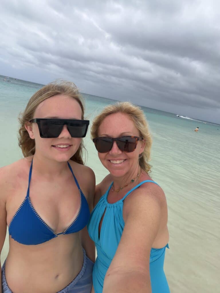 A close-up selfie of two smiling women, a teenager and an adult, standing in the clear, shallow turquoise water of a tropical beach. The younger woman on the left wears a dark blue bikini and black sunglasses, while the blonde woman on the right wears a bright teal one-piece swimsuit and tortoiseshell sunglasses. The background features a wide expanse of ocean meeting a cloudy, dramatic sky, capturing a memorable beach moment on a vacation with straight away travel.