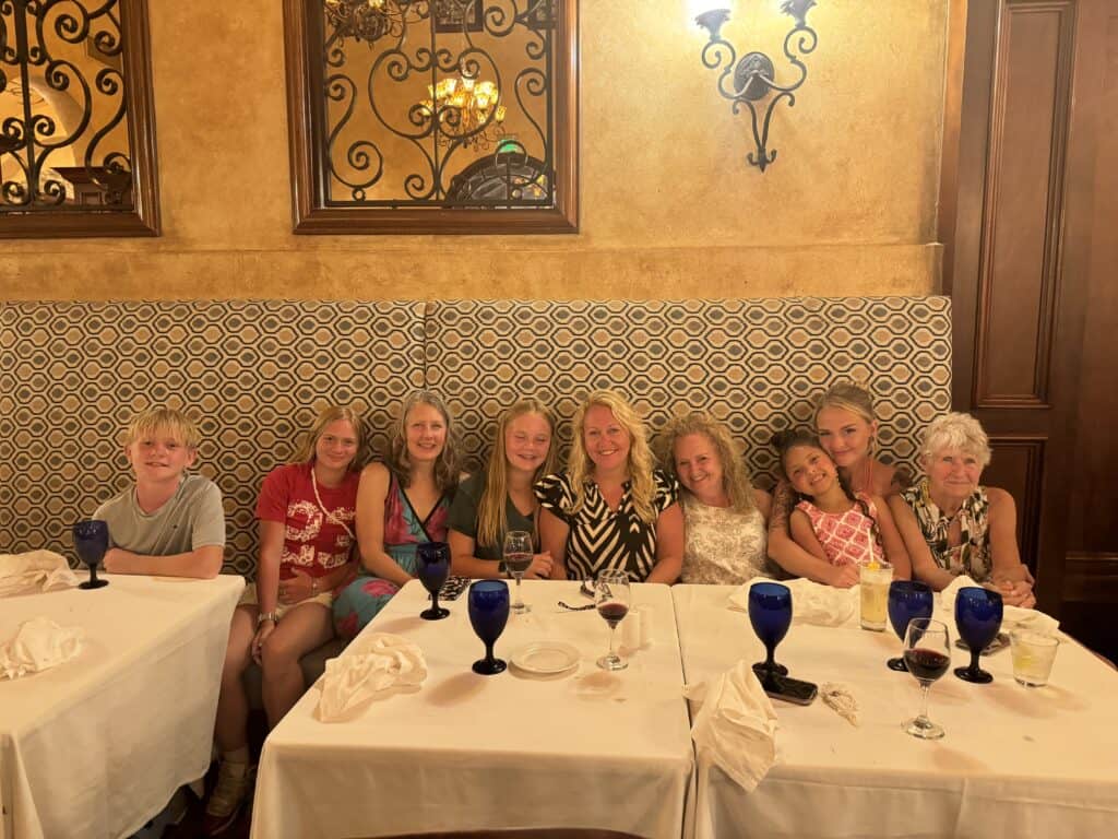 A large, multi-generational family group of nine people smiling together while seated along a long banquet booth at a restaurant. They are positioned behind tables dressed in white tablecloths, which are set with blue water goblets, wine glasses, and white napkins. The restaurant features a warm, upscale ambiance with textured golden walls, decorative wrought-iron wall grilles, and soft lighting, capturing a special dining experience shared during a straight away travel group itinerary.