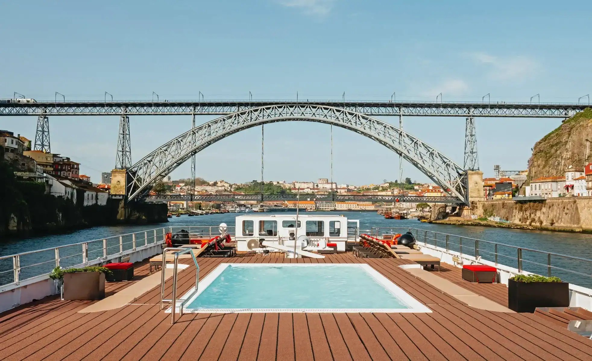 A view from the wooden sun deck of a river cruise ship, featuring a small, square swimming pool with clear blue water in the foreground. In the background, the ship is approaching a massive, historic double-deck metal arch bridge that spans across a wide river. A sunlit European city with terracotta-roofed buildings is visible along the riverbanks under a pale blue sky, offering an ideal itinerary option for straight away travel.