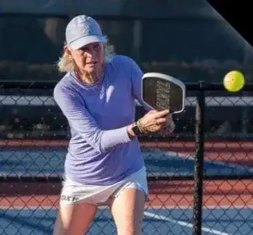 A representative from Straight Away Travel focuses intently on an outdoor pickleball court as she prepares to swing her paddle. She is wearing a light purple, long-sleeved athletic shirt, a white tennis skirt, and a grey baseball cap, with her hands gripping the paddle firmly behind a chain-link fence on a sunny day.