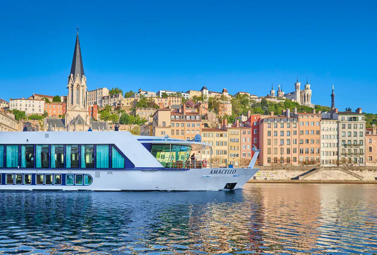 A sleek white river cruise ship gliding smoothly across calm blue waters, reflecting the vibrant city behind it. The background features a picturesque European hillside city with colorful, densely packed buildings along the waterfront, a historic church spire rising on the left, and a hilltop basilica on the right under a clear, bright blue sky.  framing a scenic route perfect for straight away travel.