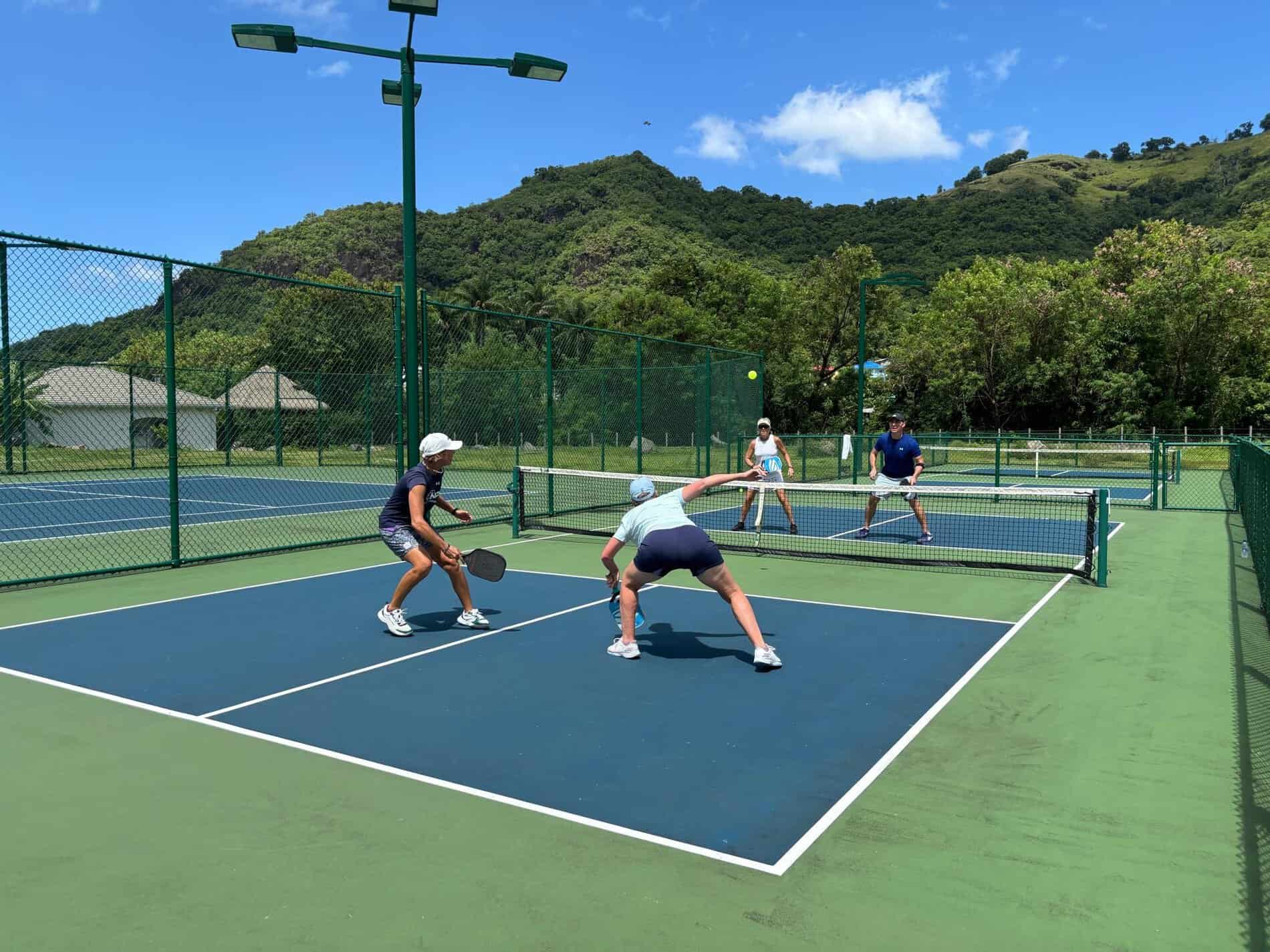 An active travel experience captured by Straight Away Travel shows a lively doubles pickleball match taking place on an outdoor court. Four players are positioned on a blue and green court surface, with one player in the foreground lunging forward to return a shot. The court is surrounded by a green chain-link fence, set against a beautiful tropical backdrop of dense green trees and a large, rolling green hill under a clear blue sky with a few light clouds.
