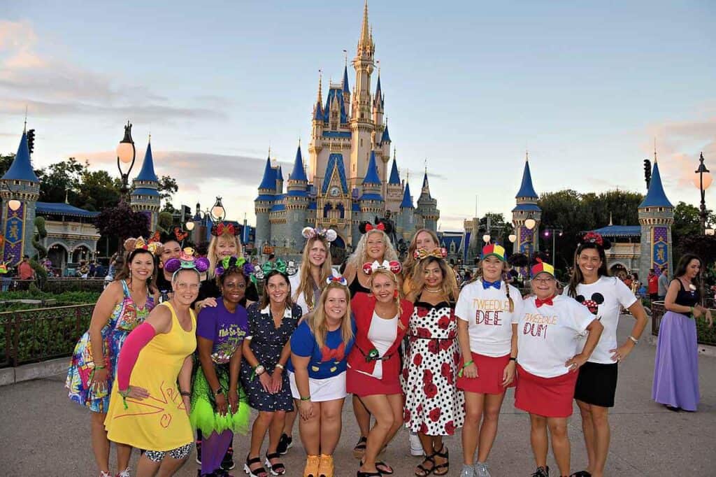 A group of travel advisors representing Straight Away Travel poses happily together in front of Cinderella Castle at Walt Disney World. The group is dressed in festive, Disney-themed outfits, with many wearing Mickey or Minnie Mouse ear headbands and colorful skirts. The iconic fairy-tale castle towers in the background under a soft, late-afternoon sky.