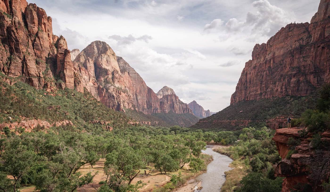 An advertisement for Straight Away Travel showing a scenic, wide-angle view of a valley floor covered with lush green trees. A winding river flows along the right side of the valley, framed by massive, steep red rock canyon walls rising dramatically on both sides under a partly cloudy sky.