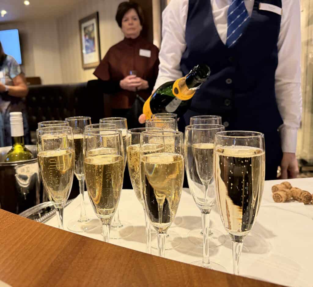 A close-up shot of an upscale reception or tasting event on a Straight Away Travel vacation. A server wearing a white shirt, blue vest, and striped tie is pouring sparkling wine or champagne from a dark green bottle into a collection of filled glass flutes arranged on a white tablecloth. In the blurred background, a female guest watches the service inside a warmly lit lounge.