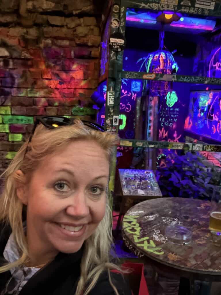 A close-up selfie of a smiling blonde woman traveling with Straight Away Travel, captured inside a vibrant, eclectic bar or club. She has sunglasses resting on top of her head. The background features an edgy, artistic atmosphere with an exposed brick wall lit by colorful lights, graffiti-covered surfaces, neon decorations, and an old-school arcade machine glowing in blue and purple hues. A wooden table with a glass is visible to the right.