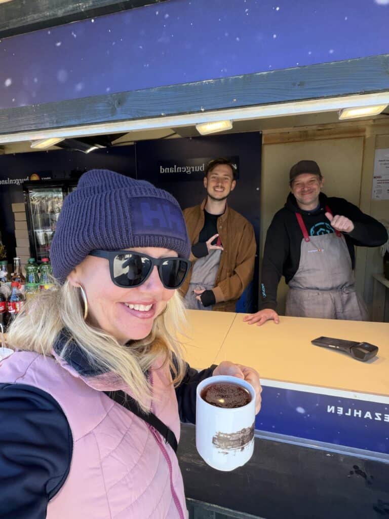 A close-up selfie of a smiling blonde woman on a Straight Away Travel vacation, holding a white mug filled with a dark beverage in the foreground. She is dressed for cool weather in a pink quilted vest, sunglasses, and a dark blue knit beanie. In the background, two friendly male vendors stand behind a counter under a wooden stall structure, smiling and gesturing warmly toward the camera.