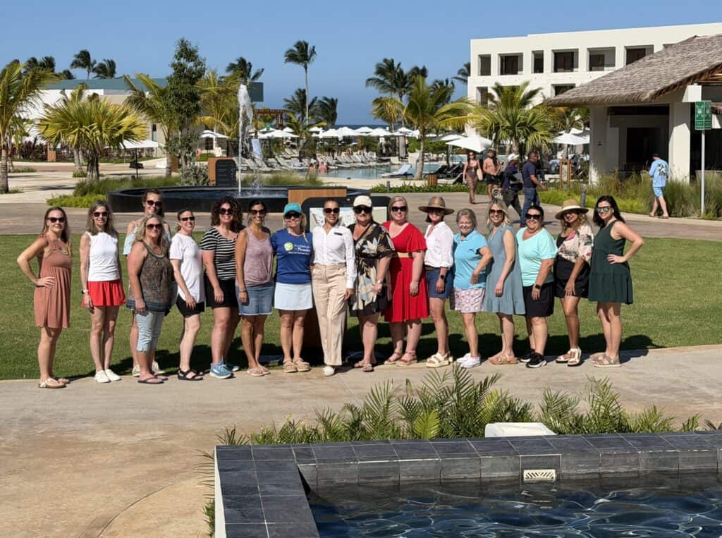 A large group of travel advisors from Straight Away Travel stands together for a group photo at a tropical resort. The women are dressed in casual summer attire, dresses, and sunglasses, lined up on a walkway next to a grassy lawn. In the sunny background, there are palm trees, resort buildings, lounge chairs, a fountain, and a glimpse of the ocean under a clear blue sky.