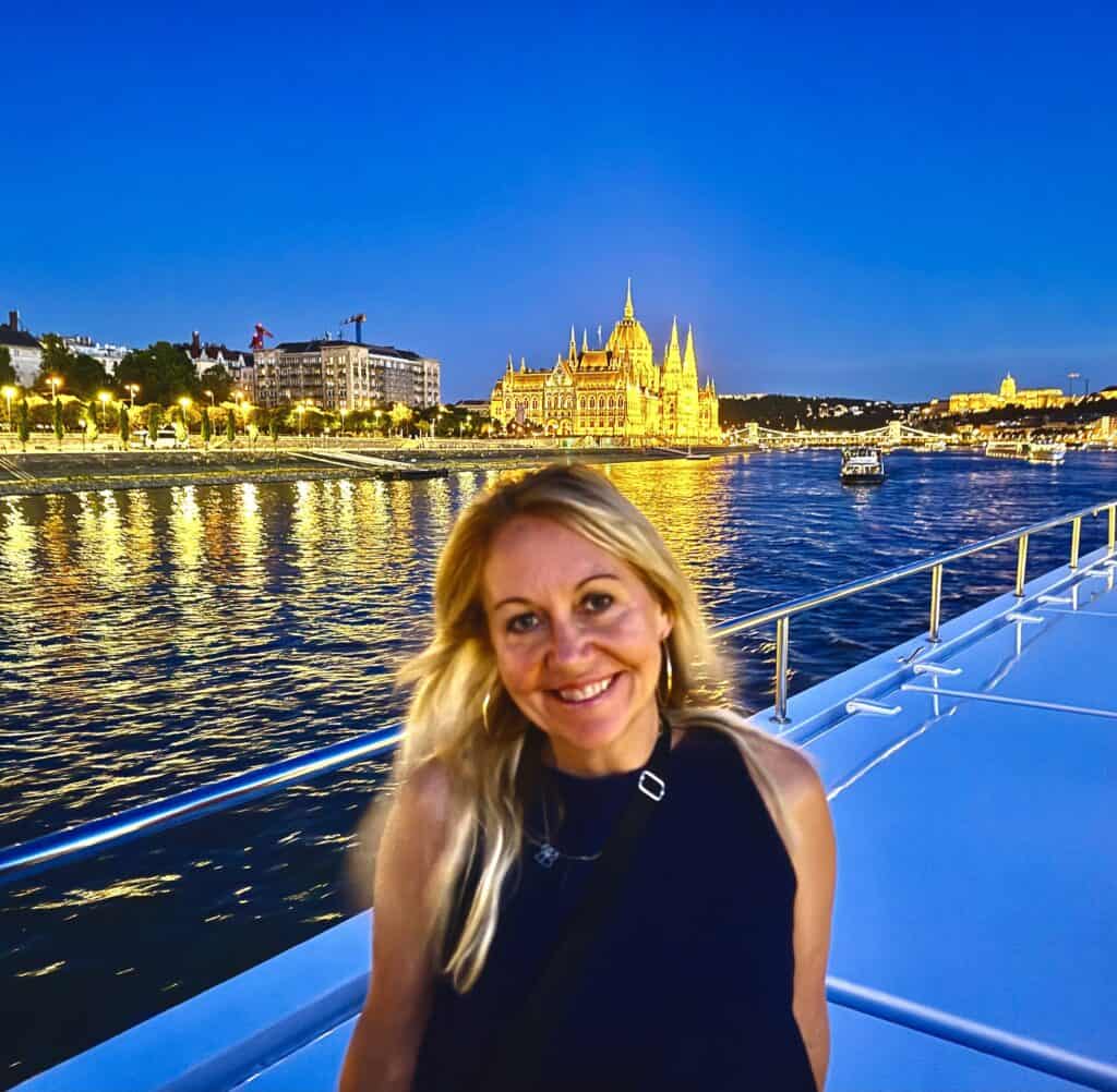 A smiling blonde woman on a Straight Away Travel vacation poses on the deck of a boat during a twilight cruise. She is wearing a sleeveless dark top and hoop earrings. In the background, the Hungarian Parliament Building in Budapest is brilliantly illuminated in golden light, with its reflection shimmering across the dark blue waters of the Danube River under a deep blue evening sky.