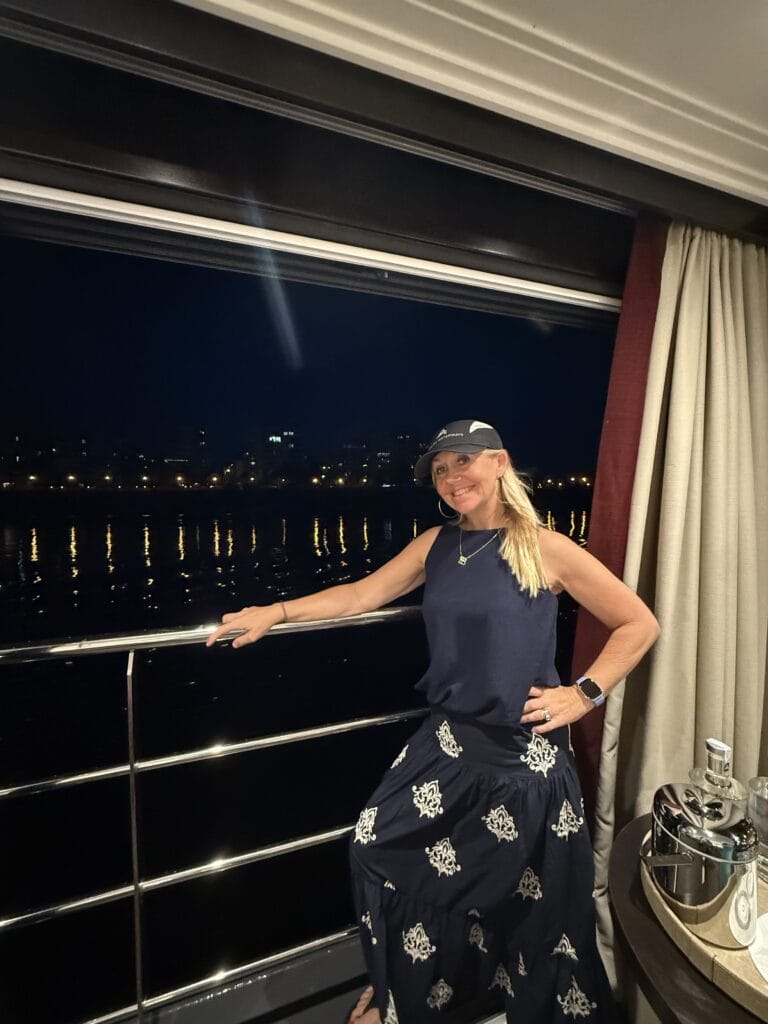 A smiling blonde woman on a cruise vacation organized by Straight Away Travel poses at night on a ship balcony. She is wearing a sleeveless dark blue top, a matching long skirt with white embroidered patterns, and a dark baseball cap. She rests one hand on the silver balcony railing, with the dark water and a glowing city skyline reflecting across the river or sea behind her.