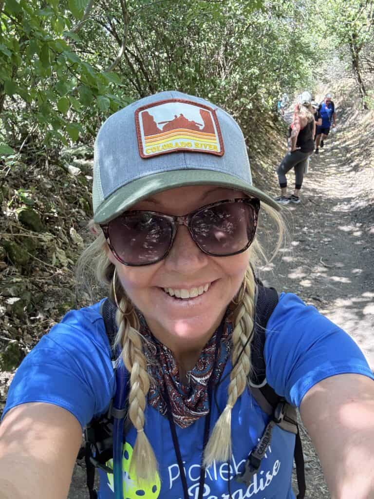 A close-up selfie of a smiling woman with blonde braided hair hiking on a sun-dappled trail, wearing sunglasses and a grey and green trucker hat featuring a "Colorado River" patch. She is wearing a blue t-shirt with a "Pickleball in Paradise" logo from the company Straight Away Travel. Other hikers are visible on the narrow, dirt path behind her, surrounded by dense green trees and bushes.