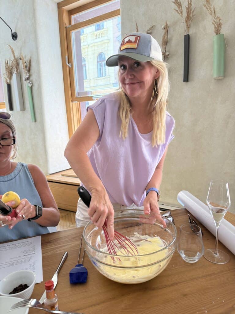 A blonde woman wearing a light purple cap-sleeve top and the same grey and green trucker hat from Straight Away Travel stands over a wooden table, smiling at the camera while whisking a yellow mixture in a large glass bowl with a red whisk. To her left, another woman in a blue top sits at the table handling a lemon, with a printed document and a small bowl of ingredients nearby. The bright indoor setting features minimalist decor with narrow wall vases holding dried grass.