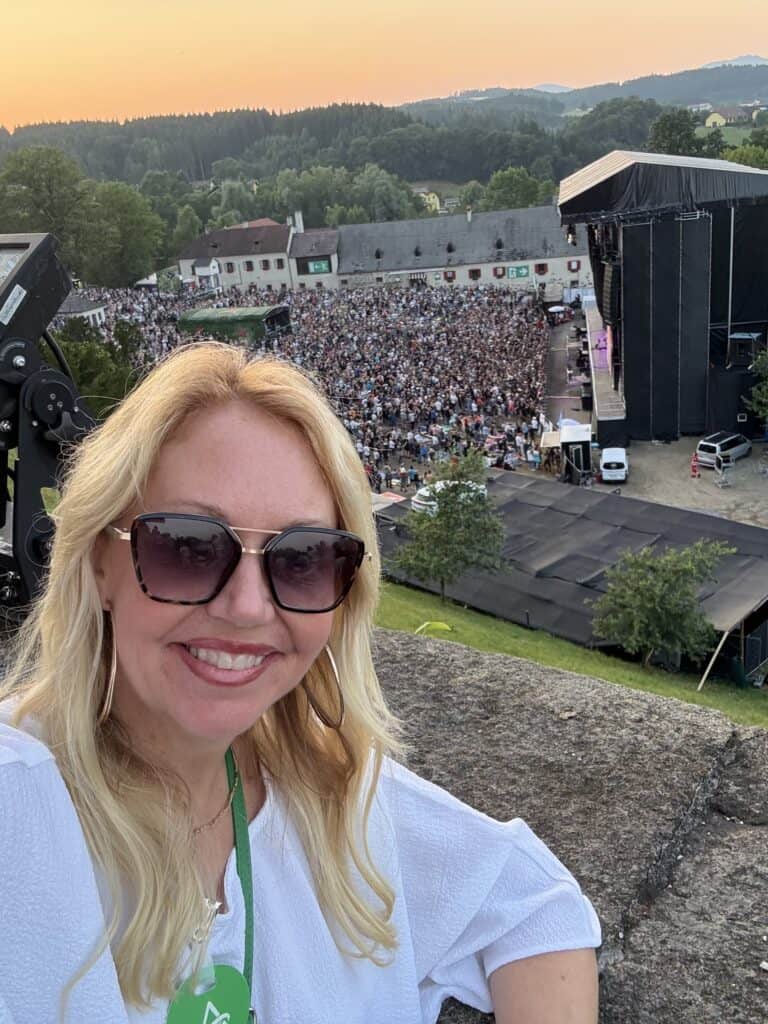An outdoor event experience featured by Straight Away Travel, showing a close-up selfie of a smiling blonde woman wearing oversized sunglasses and a white top. In the background below her, a large crowd gathers for an open-air concert in front of a massive black stage structure, set against a backdrop of historic stone buildings, rolling green hills, and a warm sunset sky.