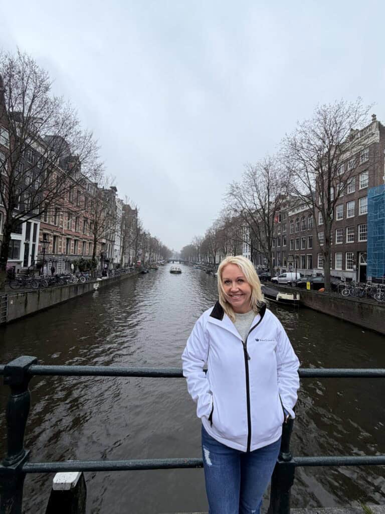 A woman representing Straight Away Travel stands on a bridge overlooking a scenic canal in Amsterdam. She is smiling, wearing a white zip-up jacket with a small logo and blue jeans, with her hands in her pockets. The canal stretches into the background, lined with traditional narrow European buildings and bare trees under an overcast sky.