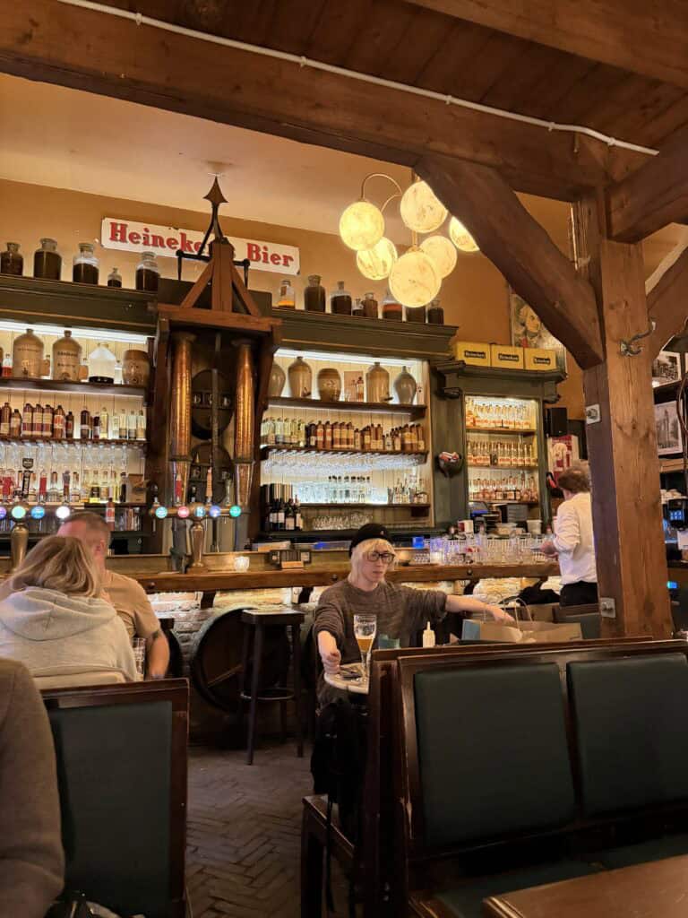 A representative from Straight Away Travel enjoys a drink inside a cozy, traditional European tavern. The interior features warm wooden beams, a large bar fully stocked with various bottles and glassware, and a vintage "Heineken Bier" sign. Patrons are seated at tables and along the bar, creating a relaxed, authentic local atmosphere.