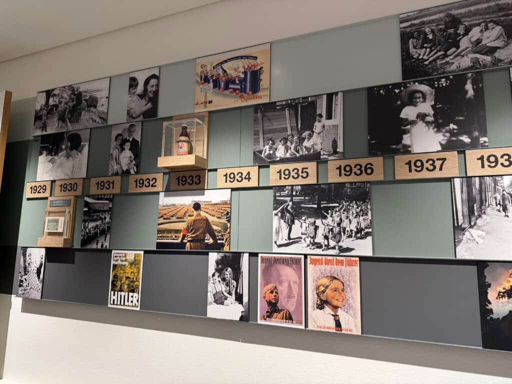 A historical exhibition display, documented by Straight Away Travel, features a chronological timeline tracking the years 1929 through 1937. The wall-mounted exhibit includes black-and-white photographs of everyday life, family portraits, and children, alongside archival Nazi propaganda posters, a labeled bottle in a glass display case, and images of historical events from the pre-WWII era.