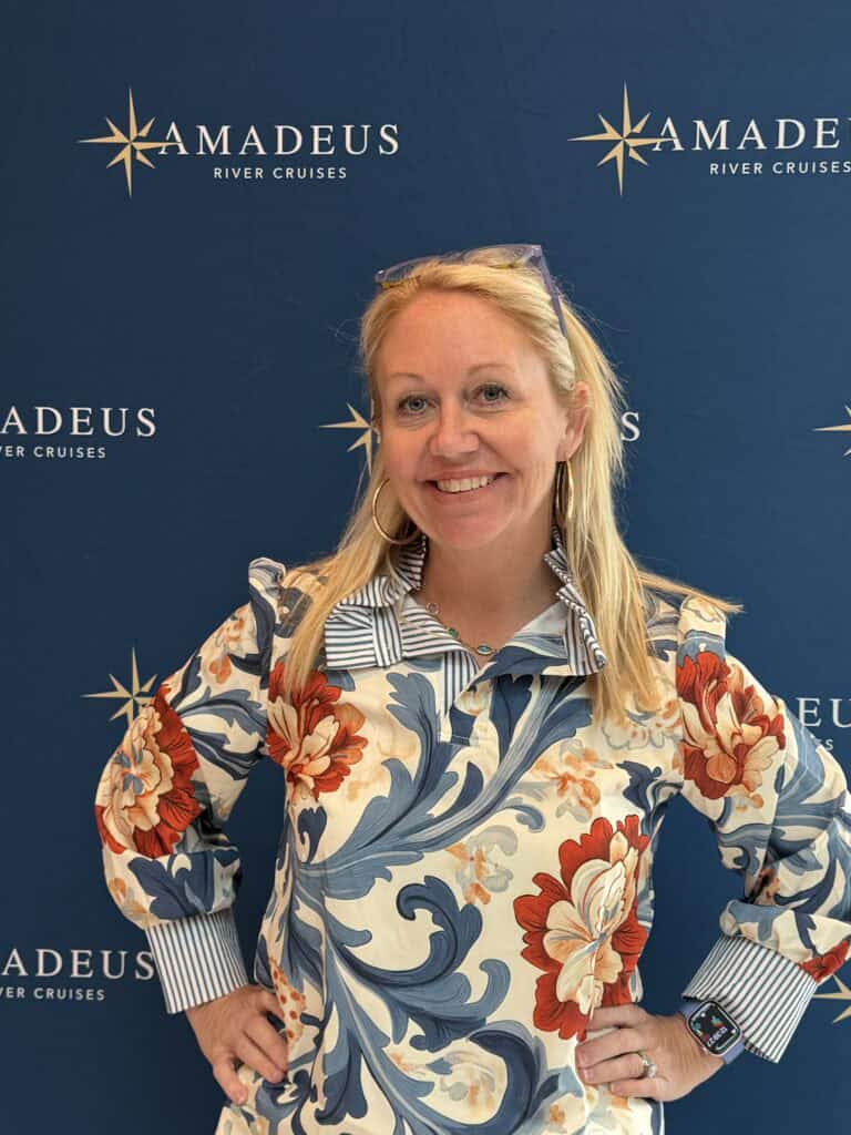 A smiling blonde woman on a Straight Away Travel vacation poses with her hands on her hips in front of a dark blue backdrop. She is wearing a cream-colored top featuring a bold, colorful floral and blue scroll pattern with striped cuffs and collar. The backdrop is patterned with a repeating gold star logo and the text "AMADEUS RIVER CRUISES."
