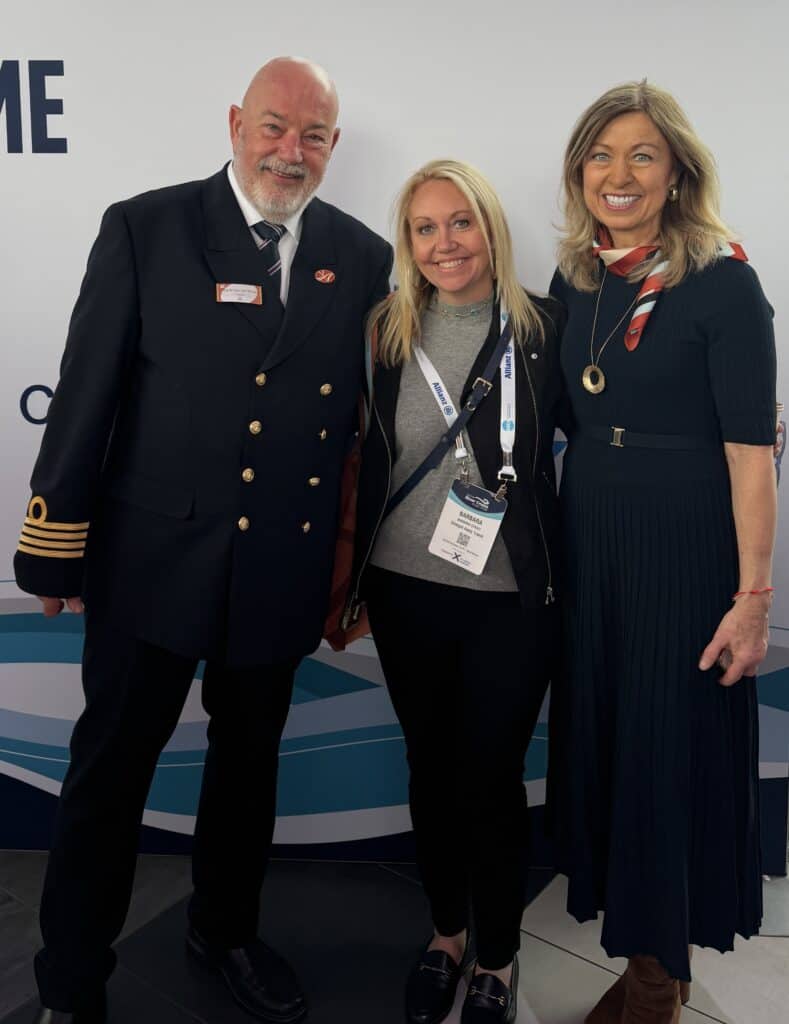 A smiling blonde woman on a Straight Away Travel vacation poses between a ship's captain and another female guest or hostess against a white and blue patterned background. The captain on the left has a white beard and is wearing a dark double-breasted dress uniform with gold sleeve stripes and a nametag. The blonde woman in the center wears black pants, a grey top, a dark jacket, and event lanyards. The woman on the right wears a dark navy pleated dress, a patterned neck scarf, and a gold necklace.