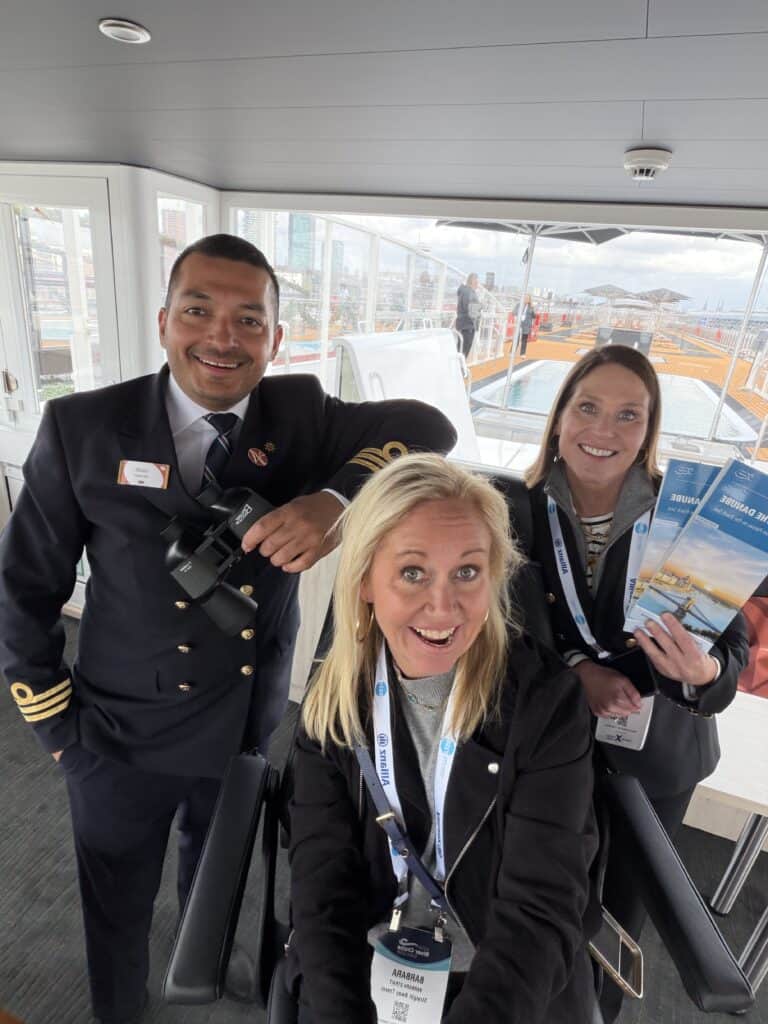 A smiling blonde woman on a Straight Away Travel cruise experience sits in the captain's chair inside a ship's bridge, looking up excitedly at the camera. Standing to her left is a ship's officer in a dark, double-breasted uniform with gold stripes on the sleeves, holding binoculars and leaning forward with a smile. To her right, another woman stands smiling while holding cruise brochures. Large windows behind them reveal the ship's outdoor deck and pool area.
