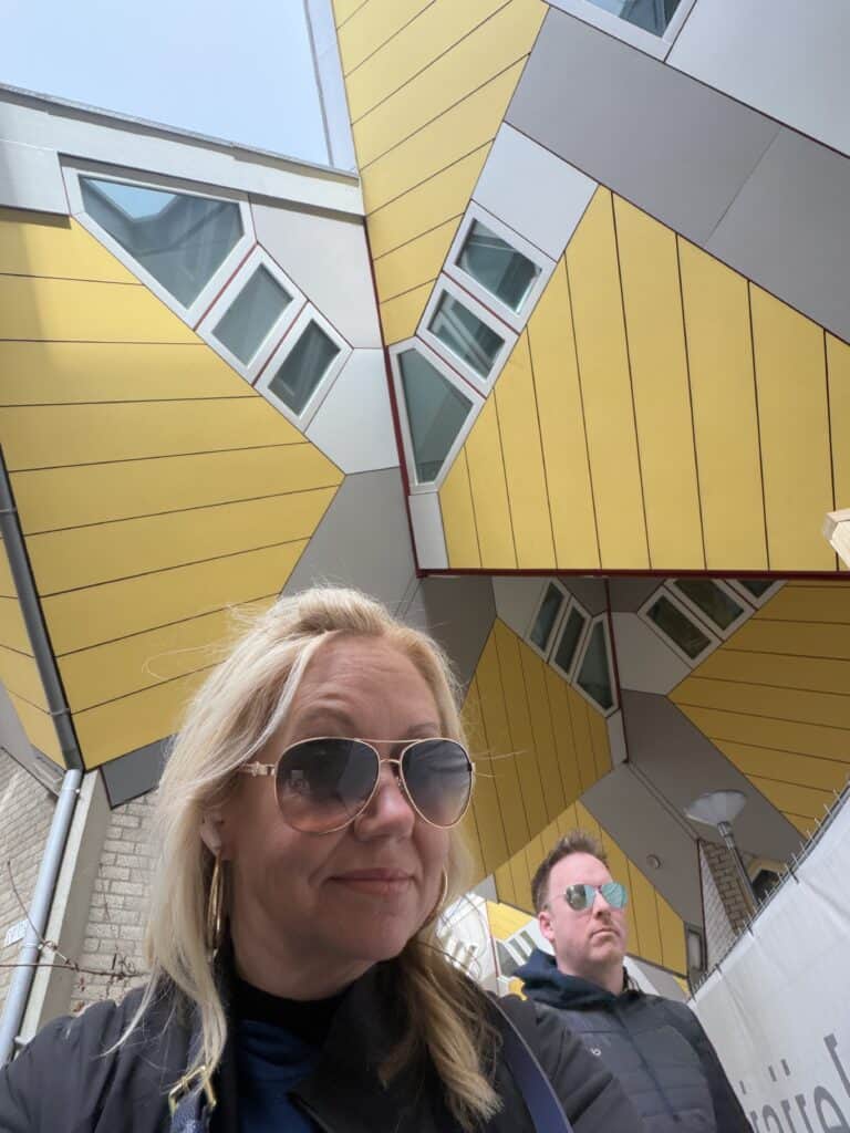 A travel experience captured by Straight Away Travel shows a low-angle selfie of a blonde woman wearing aviator sunglasses and a black jacket, with a man standing slightly behind her. They are posing in front of the famous Cube Houses in Rotterdam, showcasing the striking and unusual geometric architecture with its bright yellow and grey panels, tilted windows, and sharp angles against a pale sky.