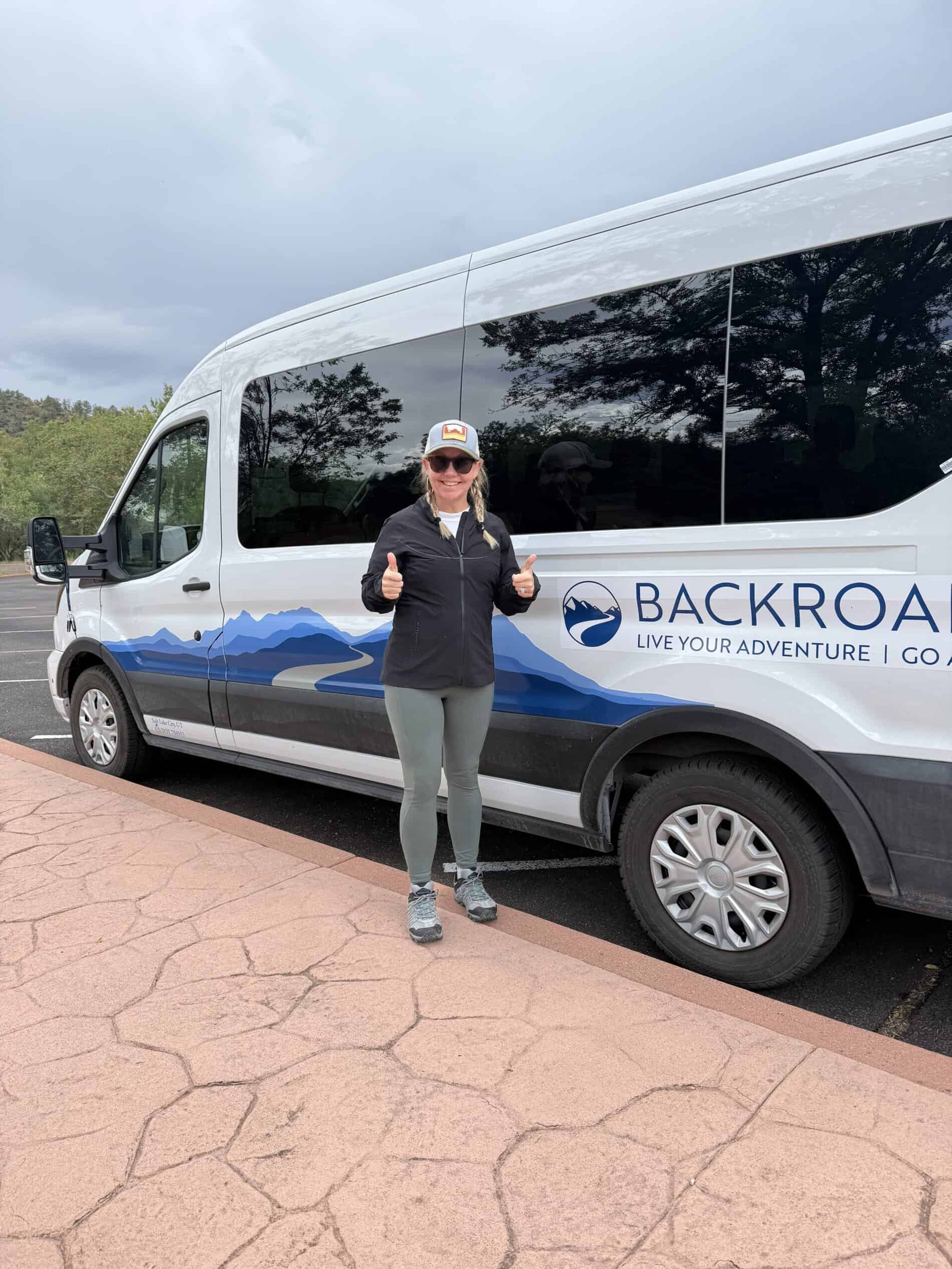 An advertisement for Straight Away Travel showing a woman smiling and giving two thumbs up while standing next to a white tour van. The van features a blue mountain graphic on the side alongside partial text that reads "BACKROADS | LIVE YOUR ADVENTURE". The woman is wearing a black jacket, grey leggings, hiking shoes, sunglasses, and a baseball cap, standing on a textured stone walkway next to an asphalt parking area.