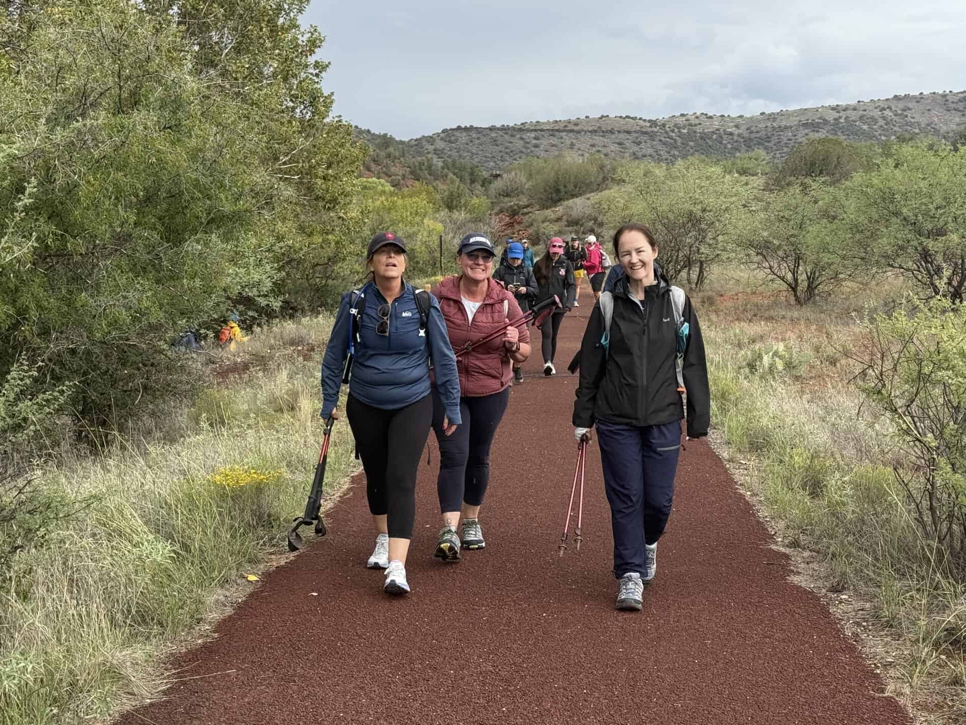 
An advertisement for Straight Away Travel showing a group of people smiling and walking down a reddish-brown gravel trail during a group hike. The hikers are dressed in jackets, activewear, and baseball caps, with some using trekking poles. The path is lined with green bushes and sparse vegetation, with low hills visible in the background under an overcast sky.