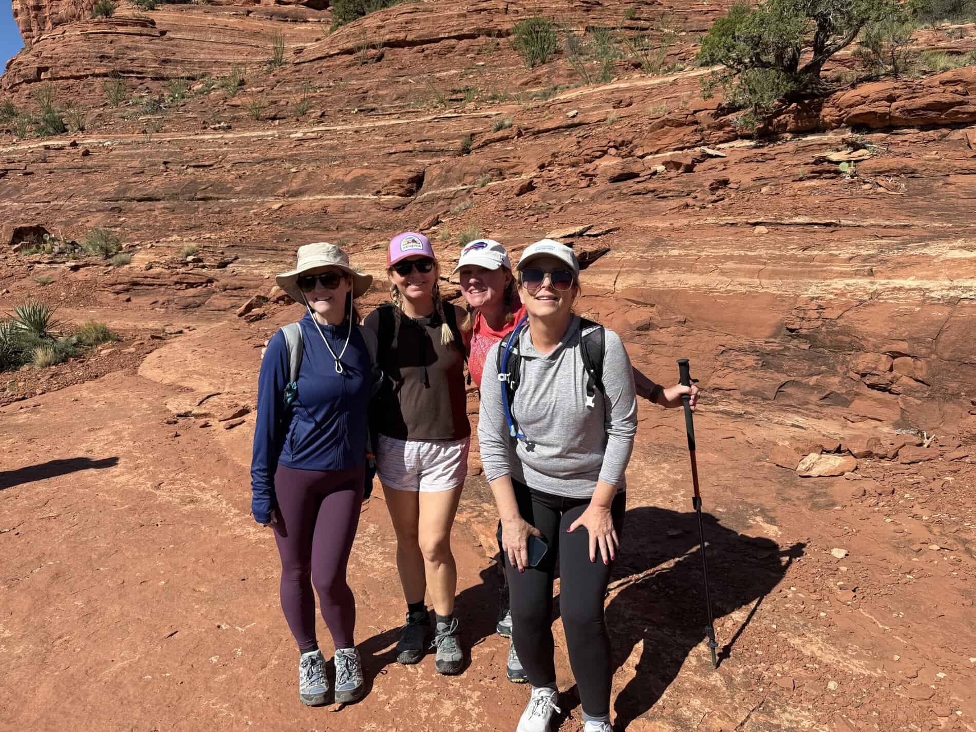 An advertisement for Straight Away Travel showing a group of four women smiling together during a hiking trip. They are wearing activewear, hats, and sunglasses, with one holding trekking poles. They are standing on a reddish dirt path with a large, textured red rock formation rising steeply in the background under bright sunlight.