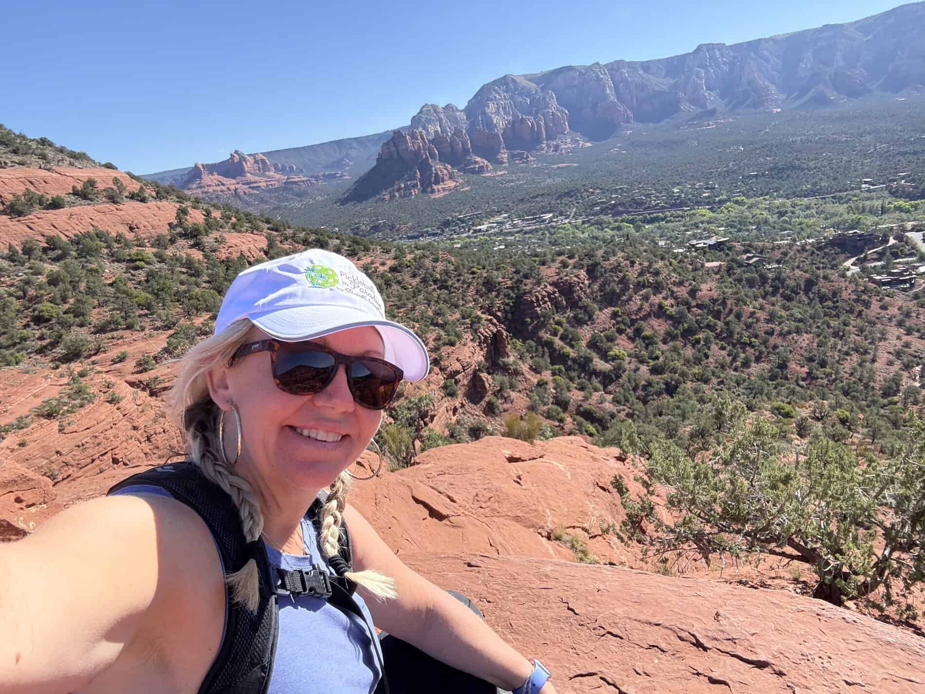 An advertisement for Straight Away Travel featuring a smiling woman taking a selfie while hiking. She is wearing sunglasses, a white baseball cap, and a backpack strap is visible. She is sitting on a red rock landscape, with a vast valley filled with green trees and dramatic red rock mountains stretching into the distance under a clear, bright blue sky.