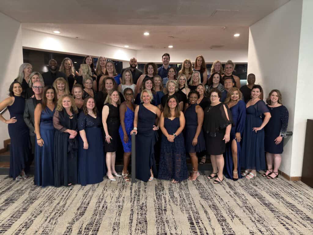 A large group of elegant professionals representing Straight Away Travel poses together for a formal group portrait indoors. The group is dressed in sophisticated evening wear, predominantly in shades of dark blue, navy, and black dresses and suits. They are arranged in multiple rows on a small set of stairs in an upscale venue, with patterned carpeting in the foreground and warm overhead lighting.